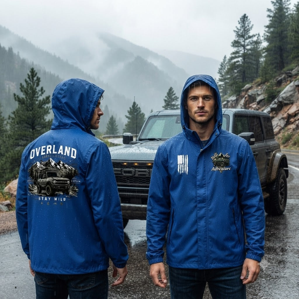 Two men wearing blue hooded jackets standing on a wet mountain road in front of a black SUV, with misty pine mountains in the background. One man faces away showing a large back graphic that reads OVERLAND and STAY WILD, and the other faces forward showing a chest logo.